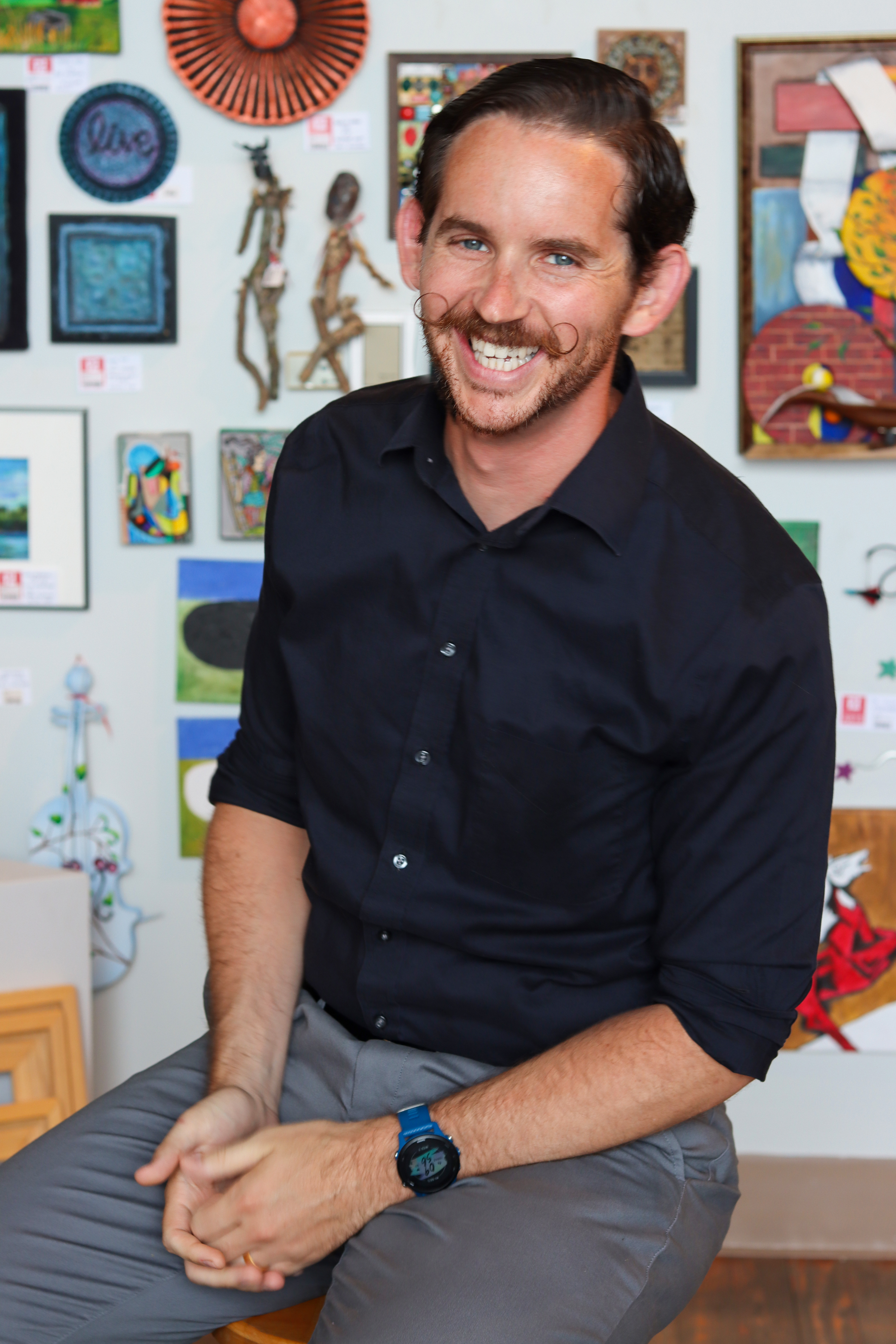 A smiling man with a mustache sitting in front of a colorful wall adorned with various art pieces. He is wearing a black shirt and gray pants.